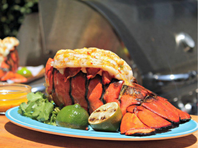Grilled lobster tail with lemons halves on a blue plate, sitting on wood table. Standup propane grill in the background.